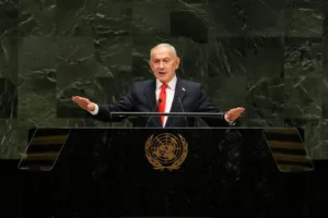 The image shows a man speaking at a podium with the United Nations emblem, indicating he is addressing the UN General Assembly.