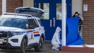 A police officer and a forensic investigator are at the scene of an incident outside a building, with a police vehicle parked nearby.