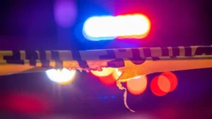 A police barricade tape is illuminated by flashing blue and red lights, indicating an active emergency scene.