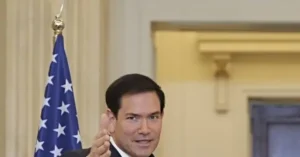 The image shows a man in a suit giving a thumbs-up gesture in front of an American flag.