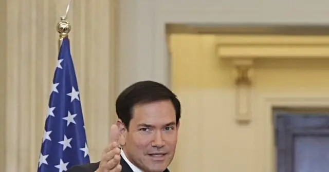 The image shows a man in a suit giving a thumbs-up gesture in front of an American flag.