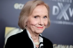 The image shows a woman with short, light-colored hair, wearing a black blazer and pearl necklace, smiling at the camera.