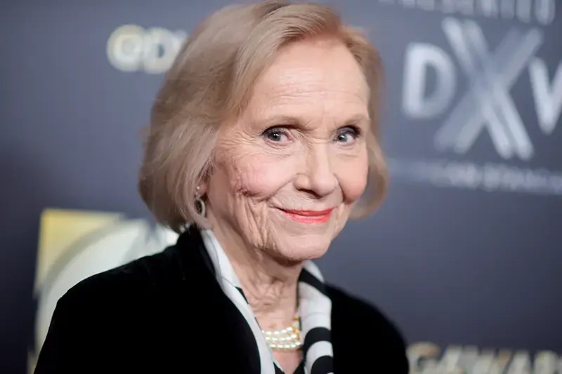 The image shows a woman with short, light-colored hair, wearing a black blazer and pearl necklace, smiling at the camera.