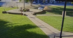 A car is seen driving through a landscaped area, causing a cloud of dust to rise.