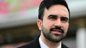 The image shows a man with a beard and short hair, dressed in a suit, standing in an outdoor setting with a blurred background.
