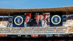 The image shows a large stadium screen displaying the Presidential Seal and a group of individuals, with a crowd of spectators in the stands.