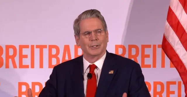 The image shows a man in a suit and tie speaking at a podium with the word "BREITBART" in the background, alongside an American flag.