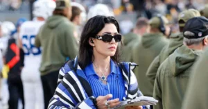 A woman in a blue and white striped jacket is seen at a football game, holding a notebook and pen, with a crowd of people and players in the background.