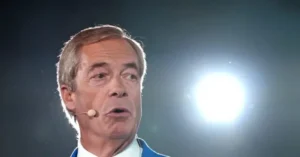 The image shows a person speaking into a microphone, with a bright light source behind them.