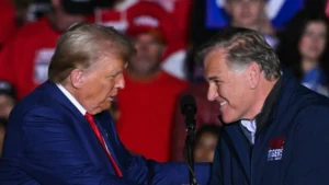 The photo shows two individuals shaking hands at a public event, with a crowd of people in the background.