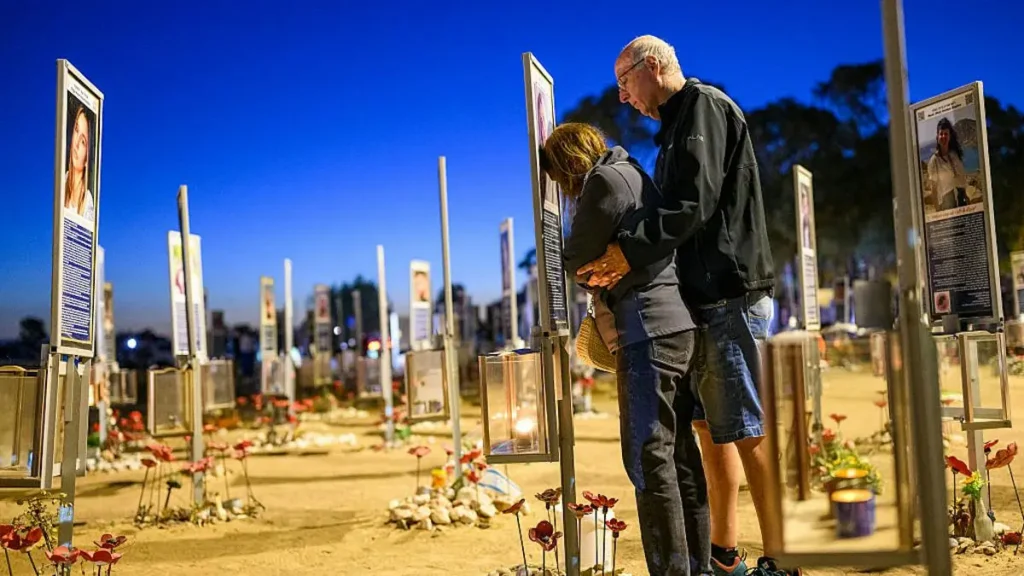 A couple embraces at a memorial site adorned with flowers and informational boards under a night sky.