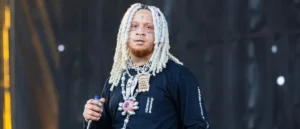 The image shows a person with long, white dreadlocks, wearing a black shirt with a pocket and a chain necklace, holding a microphone, and standing on a stage.