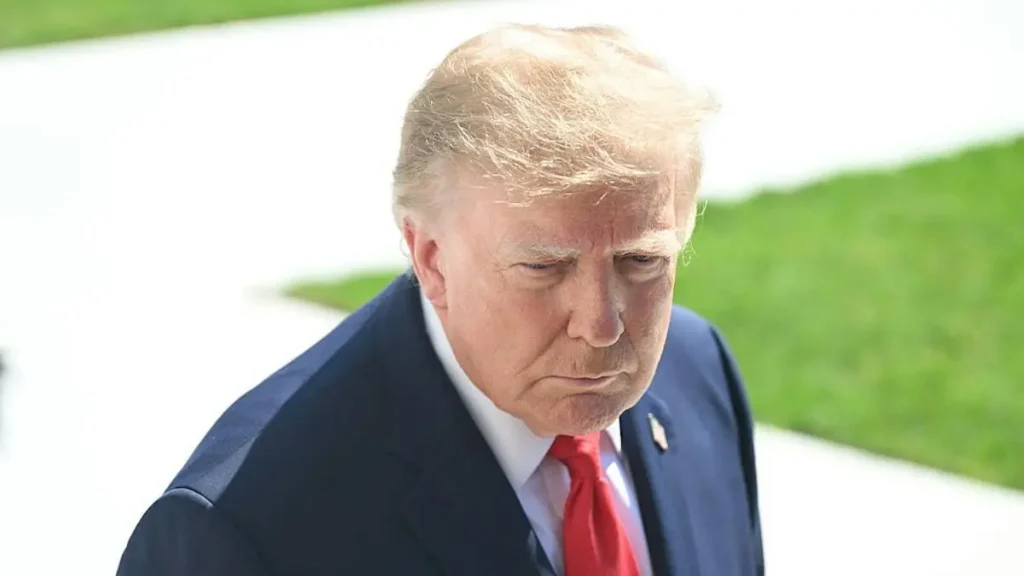 The image shows a man in a suit and red tie, looking downward with a serious expression.