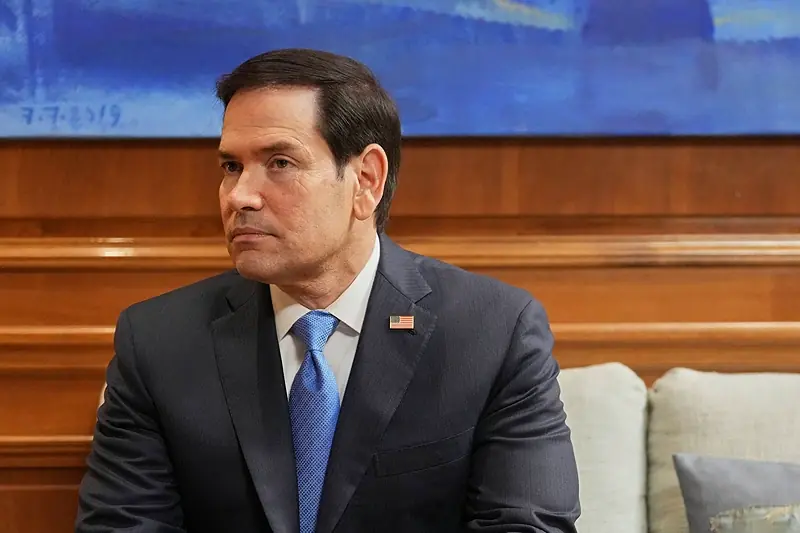 The image shows a man in a suit and tie, seated in a formal setting with a wooden panel background and a date visible in the upper left corner.