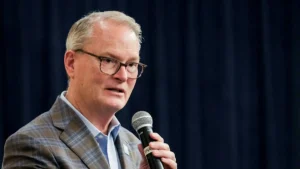 The image shows a man speaking into a microphone, dressed in a plaid blazer and glasses, against a dark blue backdrop.