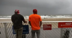 Two individuals stand on a beach observation deck, looking out at the stormy sea, with a warning sign about the drowning risk visible nearby.