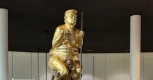 The image shows a controversial statue depicting a figure holding a scale and a stick, standing over another figure, symbolizing power and authority.