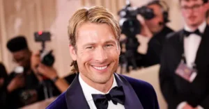 A man in a tuxedo smiles at the camera while being photographed by a group of photographers at what appears to be a formal event.