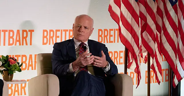 The image shows a man in a suit sitting in a chair, gesturing with his hands, with American flags and the "Breitbart" logo in the background.