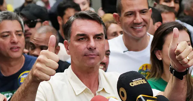 A man in a light-colored shirt gives a thumbs-up while surrounded by a crowd and microphones, suggesting a public event or press conference.