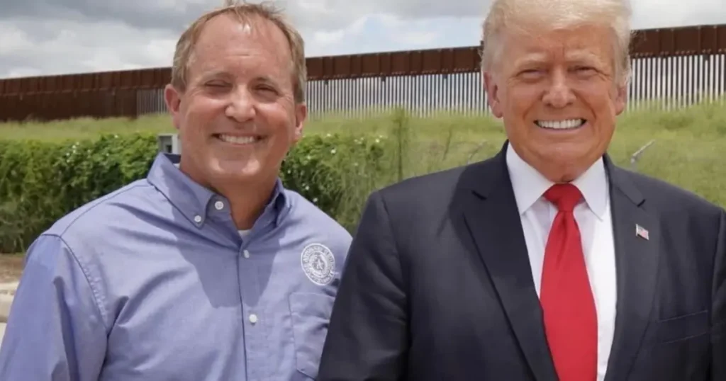 The photo shows two individuals standing in front of a border fence, with one person wearing a suit and tie and the other in a button-up shirt.