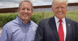 The photo shows two individuals standing in front of a border fence, with one person wearing a suit and tie and the other in a button-up shirt.