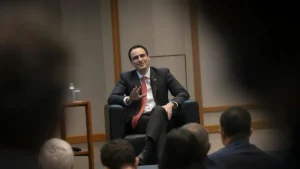 A man in a suit and tie is seated in a chair, engaging with an audience in a formal setting.