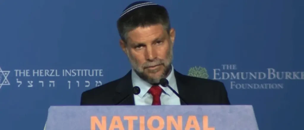 A man wearing a kippah and a suit is speaking at a podium with the words "The Herzl Institute" and "The Edmund Burke Foundation" in the background.