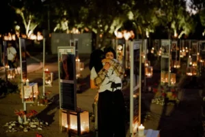 Two individuals embrace in a somber moment at a memorial site adorned with candles and photographs.
