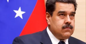 The image shows a man in a suit and tie, standing in front of a flag with a blue field and a white star, likely representing Venezuela.