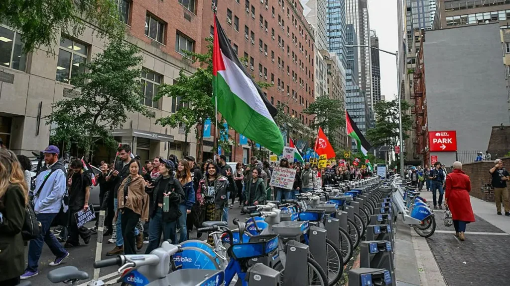 A group of people holding Palestinian flags and signs march down a city street, advocating for a cause.