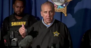 A sheriff speaks at a press conference, with a police officer standing nearby, in front of a Minnesota state emblem backdrop.