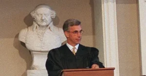 A man in formal attire stands at a podium, delivering a speech in front of a bust.