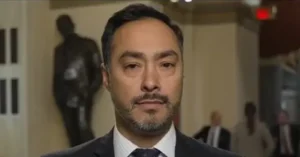 The image shows a man in a suit and tie, standing in what appears to be a formal or professional setting, possibly a news interview or press conference.