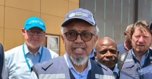 The photo shows a group of individuals, including one wearing a World Health Organization cap, standing outside a building.