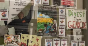 The image shows a person raising a poster in front of a wall covered with various protest signs advocating for Palestine and calling for divestment from genocide.