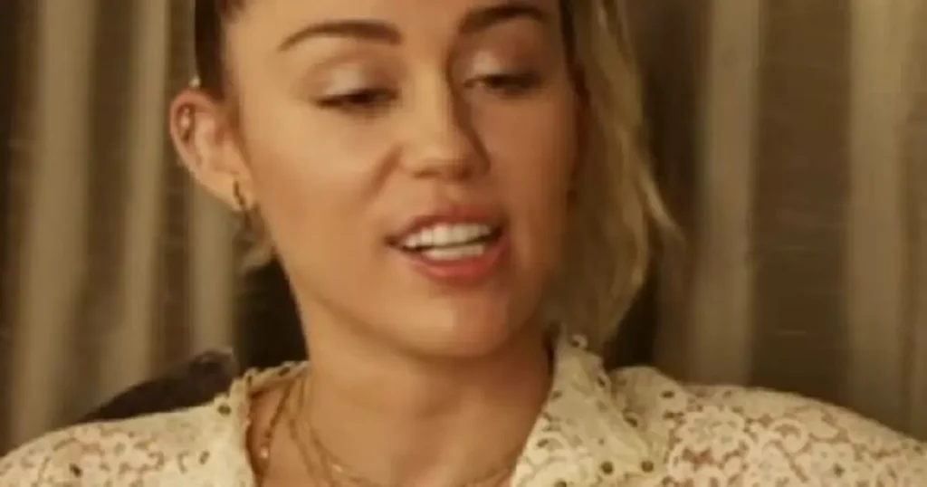 The image shows a person wearing a lace top, with a background that appears to be indoors with curtains.