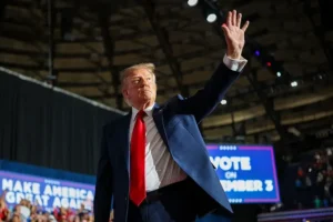 The image shows a political rally with a prominent figure waving to the crowd, accompanied by a large screen displaying campaign slogans.