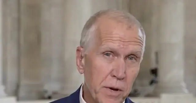 The image shows a man in a suit speaking in what appears to be a formal or official setting.