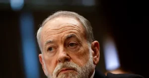 The image shows a close-up of a man with a beard and short hair, wearing a suit, in what appears to be a formal setting.