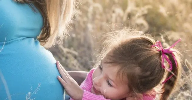 A young girl gently touches her pregnant mother's belly in a serene outdoor setting.