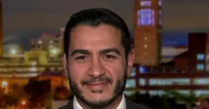 The image shows a man in a suit and tie, smiling, with a cityscape in the background.