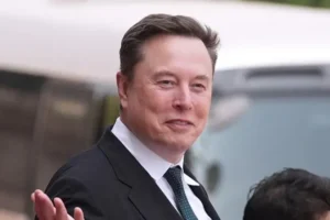 The image shows a man in a suit and tie, gesturing with his hand, with a blurred background that appears to include a vehicle.