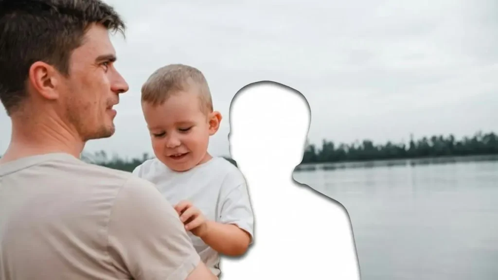 A man and a child are standing by a body of water, with the man holding the child and both looking in the same direction.