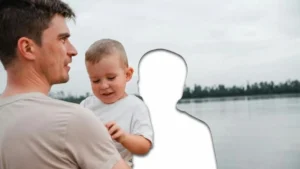 A man and a child are standing by a body of water, with the man holding the child and both looking in the same direction.