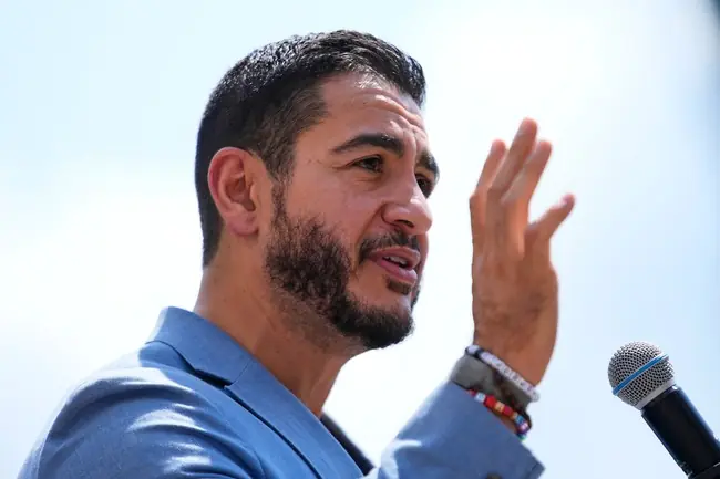 A man in a blue suit gestures with his hand while speaking into a microphone outdoors.