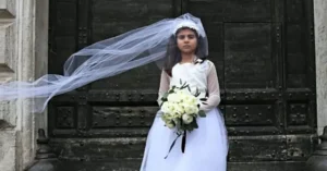 A young girl in a white dress and veil holds a bouquet of flowers, standing in front of a large, ornate door.