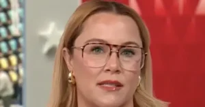 The image shows a person with long blonde hair wearing glasses and earrings, set against a red and white background.