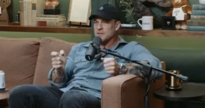 A man is sitting on a couch, speaking into a microphone in a cozy, book-lined room.