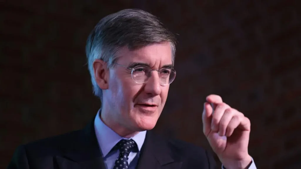 The image shows a man in a suit and tie, gesturing with his hand while speaking, suggesting he is giving a speech or presentation.